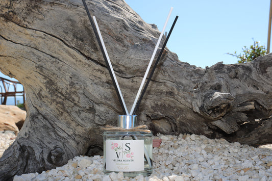 Reed diffuser with sticks in a glass bottle on pebbles with a wooden log in the background